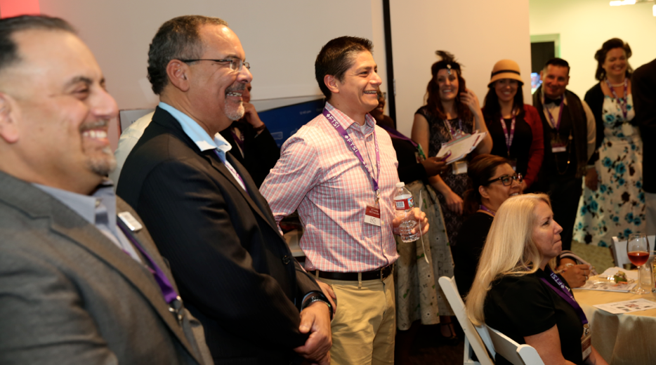 A group of happy people standing and looking to the right at a presenter. They are in business attire and wearing conference lanyards.